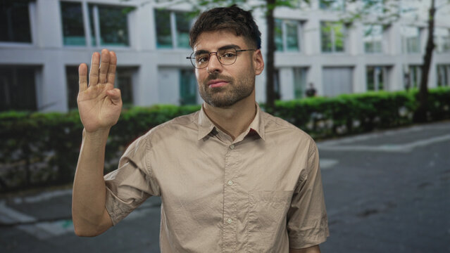 Man in beige shirt and glasses raising hand in greeting on sunlit tree lined city street; friendly greeting.
