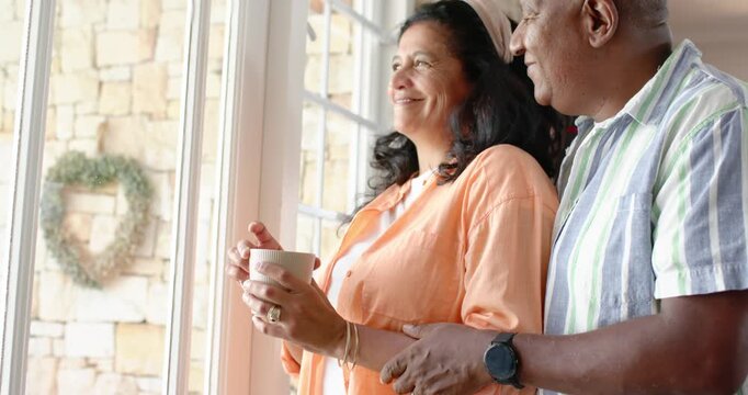 Eye contact initiating, couple are standing by paned window sharing mug and embrace watching view