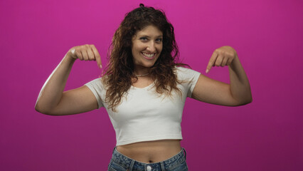 Obraz premium Woman pointing both index fingers down at her exposed navel in a magenta studio, wearing a white cropped shirt and jeans; playful confidence.