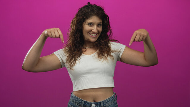 Woman pointing both index fingers down at her exposed navel in a magenta studio, wearing a white cropped shirt and jeans; playful confidence.