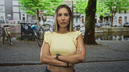 Fototapeta premium Young hispanic blonde woman with arms crossed wearing a yellow shirt on a cobbled street by a canal with parked bicycles and a large tree; determination independence.