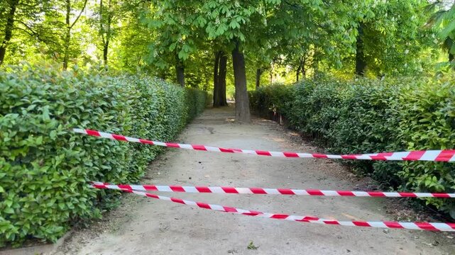 Park pathway blocked with red and white warning tape, restricted access in green garden alley, safety barrier outdoors