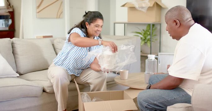 African couple opening box at home, removing bubble wrap and handing white teapot, settling home
