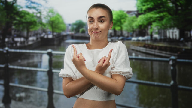 Young woman with cropped hair in a white crop top crossing arms and flipping middle fingers while smirking on a street beside a canal railing; defiance rebellion.