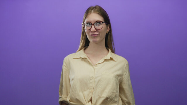 Blonde woman wearing glasses sticks out tongue, raises hands and smiles while tilting head in purple studio; mischief.