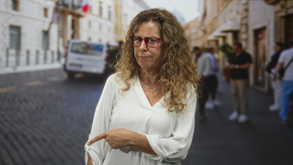 Obraz premium Woman pointing finger on street, showing hand and face with red glasses and curly hair, wearing white blouse and looking upward; curiosity travel.