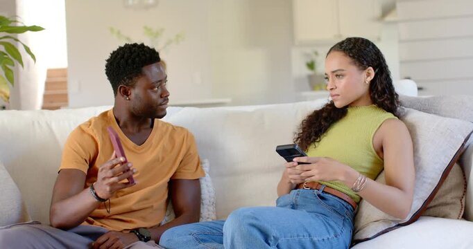 African American couple sitting on sofa, man showing pink phone after seeing message, woman tense