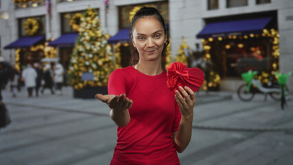 Fototapeta premium Young woman extends hand offering red heart gift box on street storefront with christmas tree; romance generosity.
