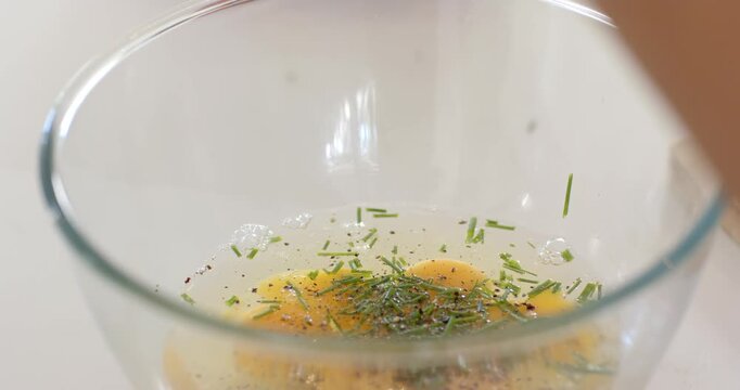 Female cook adding herbs and pepper to cracked eggs in glass bowl on countertop, preparing omelette