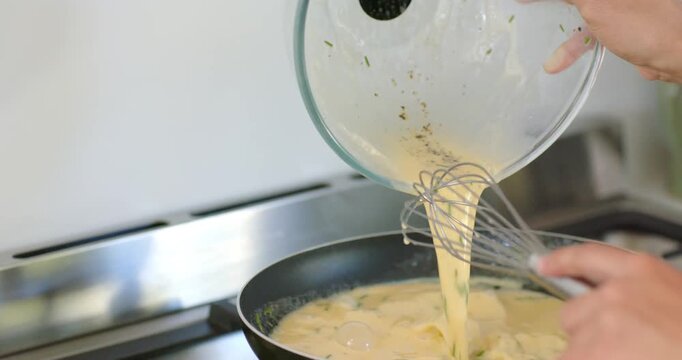Adult pouring egg mixture from glass bowl into heated pan on stove, whisking with metal whisk