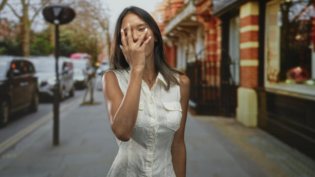 Thai woman in sleeveless white dress covering her face with her right hand on an urban city street with parked cars and a brick storefront; embarrassment vulnerability.