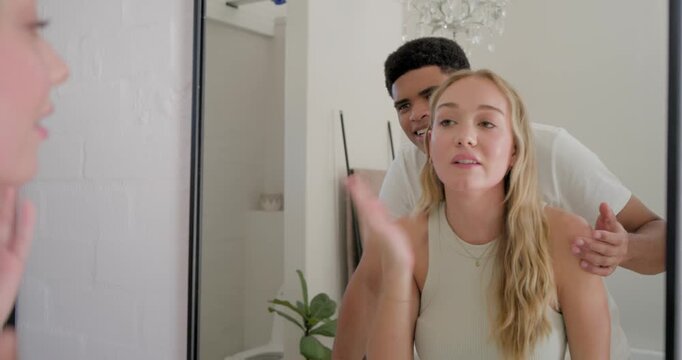 Couple at bathroom mirror, African American man pointing, woman patting face checking skin