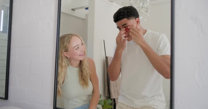 Diverse couple watching man applying bright red under-eye patches at vanity after woman pointing