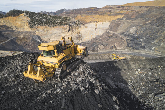 Aerial view of a large yellow bulldozer working on a pile of coal in an open-pit mine with terraced rock walls in Santa Lucia de Gordon, Castile and Leon, Spain.