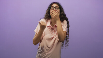 Fototapeta premium Hispanic teenage woman in pink polo covers mouth with hand while laughing and pumping fist in studio; playful amusement.
