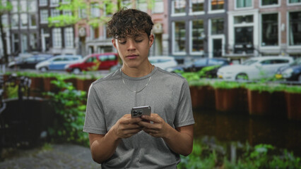 Obraz premium Teenage boy holding smartphone with fingers scrolling while looking down on a street canal by buildings; quiet thoughtful.