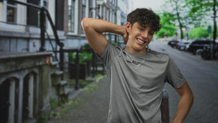 Fototapeta premium Teenage boy with hand behind neck smiling in gray t shirt on a street by a building; playful youth moment.