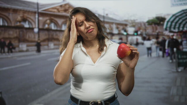 Woman holds a red apple and presses her temple with one hand in an obvious headache on a busy city street with traffic and market stalls; fatigue.