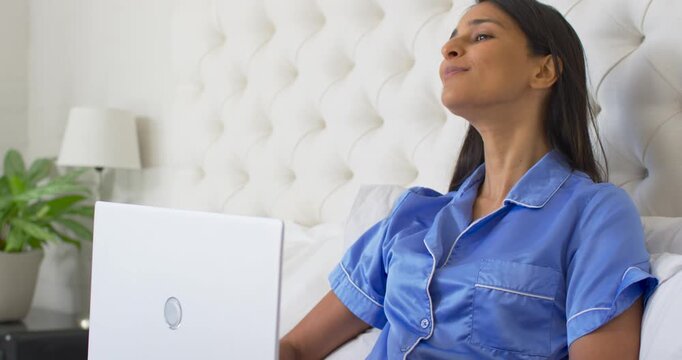Adult woman in pajamas sitting in bed seeing clip on white laptop, leaning back, returning smiling