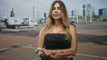 Fototapeta premium Middle age woman holding smartphone with folded arms, standing at a busy urban street intersection near a delivery van and cyclists, neutral pensive expression; contemplation.