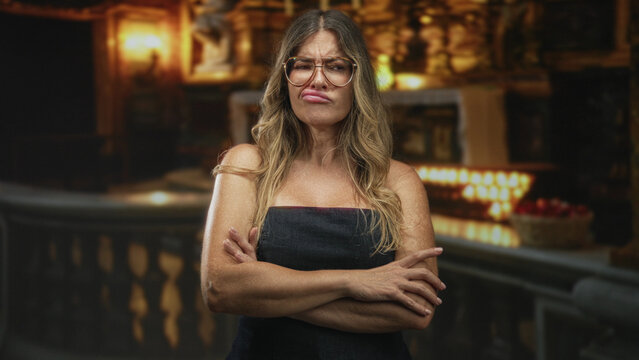 Woman with glasses and strapless dress, arms crossed and pouting lips by a balustrade in an ornate building; defiance strength.