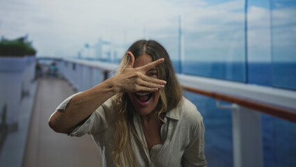 Fototapeta premium Woman covering eyes with hand on cruise ship deck, mouth open laughing, leaning by glass railing and facing sea; playful joy.