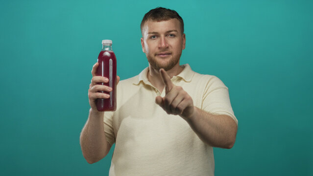 Man holding a bottle, pointing finger at the bottle and touching his chin in studio with teal backdrop; curiosity.