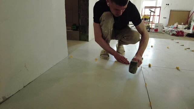 A worker lays tiles on a floor while focusing on precise placement. Tools and materials are spread around the construction site in the afternoon. The task requires careful attention.
