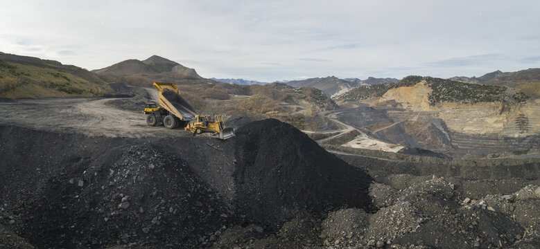 Aerial view of an open-pit coal mine with heavy machinery working on dark piles of earth in the rugged mountains of Santa Lucia de Gordon, Castile and Leon, Spain.