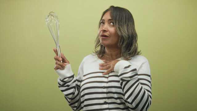 Woman holding whisk with hand on chest, striped sweater visible, expressive gasp gesture in studio; surprise.
