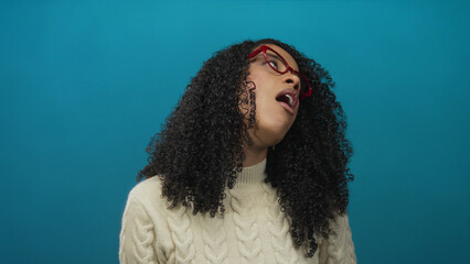 African american woman yawns with open mouth and red glasses wearing cream sweater in blue studio  fatigue. © Krakenimages.com
