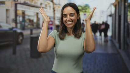 Obraz premium Woman raising hands and laughing on a bustling urban street, green shirt visible, palms and arms shown, blurred storefronts, bollards, a bin and passing pedestrians in frame; joy celebration.