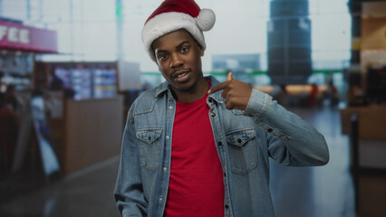 Obraz premium Young african american man wearing santa hat and denim jacket points finger to shirt at airport terminal; pride.