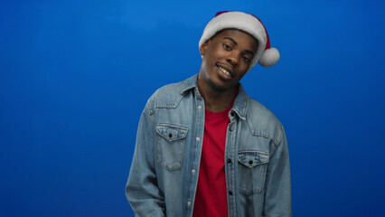 Obraz premium Man wearing santa hat, red shirt, and casual denim jacket smiles and tilts head in studio; holiday joy.