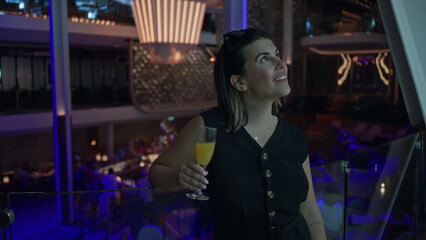 Obraz premium Young woman holds a champagne flute and sips orange drink on a glass balcony inside a building; celebration night out.