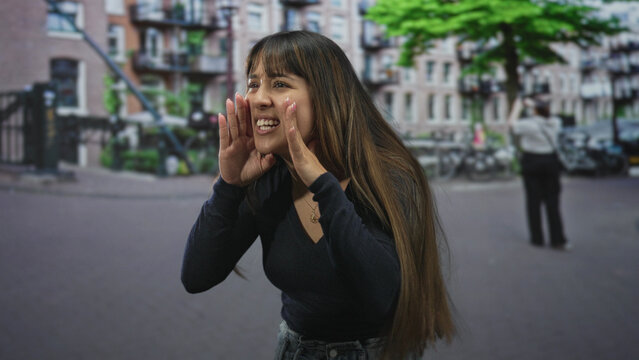 Young woman cupping hand to mouth, calling out with long hair and open palm gesture on street; spontaneous playful.