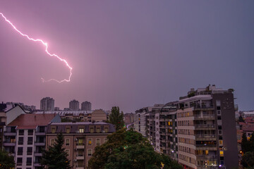 Dramatic pink lightning bolt striking across a purple stormy sky above a modern cityscape with illuminated apartment buildings during twilight. © Victor