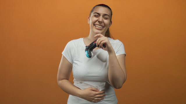 Woman in white t shirt holding a gamepad in right hand, left hand on stomach while laughing against orange studio backdrop, midgame reaction; joy play.