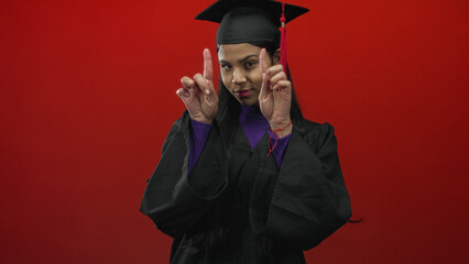 Obraz premium Young latina graduate woman in black mortarboard and gown puckers lips gesture in studio with vivid red background; pride.