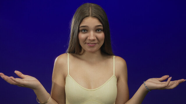 Teenage girl with bare shoulders shrugs palms up against a deep blue studio wall showing puzzled stance; uncertainty.