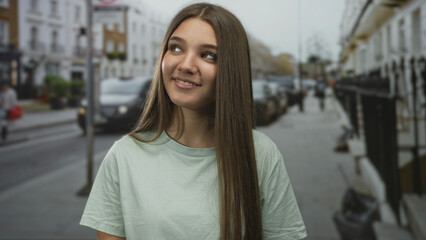 Obraz premium Girl showing smiling face with long brown hair in a mint green tshirt looking to side on city street; serenity.