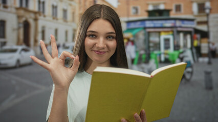 Fototapeta premium Teenage girl reading a yellow book on a city street, holding the open book with both hands at chest level; quiet concentration.