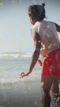 Vertical video: Girl skipping shore, wave causing splash, bubbles bokeh accenting play for travel