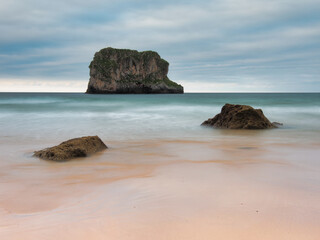 Obraz premium Coastal Sea Stack Monolith in Smooth Ocean - Long-exposure shot of a grass-topped sea stack monolith rising from the Atlantic Ocean, framed by smooth blurred water and rugged foreground rocks.
