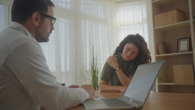 Female patient complaining of neck and shoulder pain in medical office while doctor listens and examines