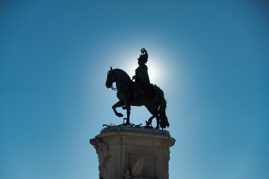 Dramatic low-angle silhouette of the bronze equestrian statue of King Jose I in Lisbon's Pra&ccedil;a do Com&eacute;rcio, featuring a striking sunburst against a bright blue sky.