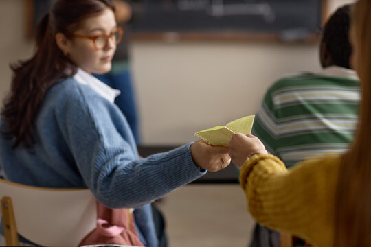 Teenage girl passing handwritten note to classmate during lesson, suggesting classroom distraction and peer communication. Useful for education campaigns, school behavior content
