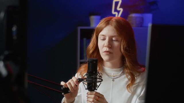 Close up of young Caucasian woman setting up professional microphone and putting on wireless headphones while sitting at computer desk and preparing for stream