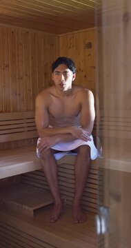 Breathing adult man sitting on wooden bench in sauna, towel wrapped around waist, glass door