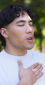 Vertical video: Asian man placing right hand on chest, breathing and opening eyes in park
